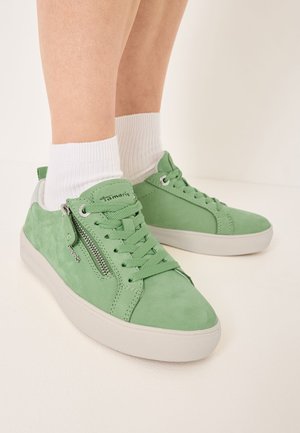 Pieds portant des baskets en nubuck vert clair avec des semelles blanches et des zippers sur les côtés, assortis à des chaussettes blanches à la cheville sur un fond neutre.