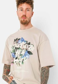 Beige katoenen T-shirt met korte mouwen, voorzien van een bloem- en vogelafbeelding in blauw en groen, en witte tekst met de tekst "Romantiseer Je Leven."