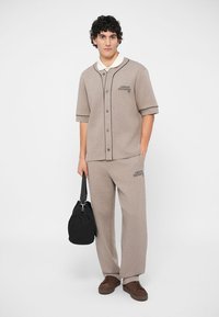 Gaiši brūns divdaļīgs apģērbs, kas sastāv no īsām piedurknēm ar pogām, kuram ir melni akcenti, un atbilstošām biksēm. Modelis tur melnu somu.