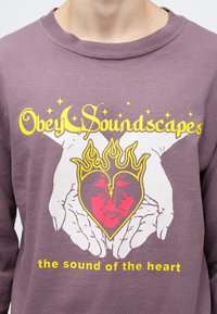 Violetinis megztinis su grafiniu dizainu, kuriame vaizduojamos rankos laikydamos širdį su veidu, geltonas tekstas viršuje ir apačioje ryškiu šriftu.