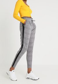 Femme portant un haut court à manches longues jaune, un pantalon écossais gris avec une bande noire sur le côté, et des baskets blanches, debout avec les mains dans les poches.