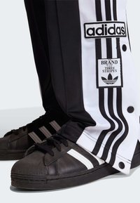 Crne i bijele Adidas trenirka hlače s tri pruge i kopčama na drukere, uparene s crnim tenisicama s bijelim prugama i gumenim potplatom.