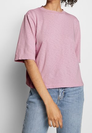 Person trägt ein locker sitzendes, hellrosa Kurzarmshirt in Kombination mit hellblauen Jeans vor einem schlichten weißen Hintergrund.