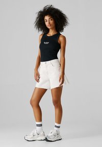 Νεαρή γυναίκα με σγουρά μαλλιά που φοράει μαύρο αμάνικο τοπ Red Bull Racing, λευκά τζιν σόρτς, λευκές κάλτσες και λευκά αθλητικά παπούτσια.