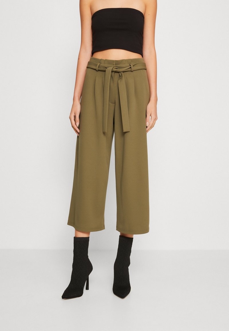 Pantalons larges vert olive, avec une ceinture nouée à la taille. Portés avec un top noir sans bretelles et des bottines noires texturées.