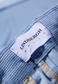 Ljusblått tyg med ett strukturerat mönster. En etikett med texten "LINDBERGH WHITE" i svart. Sömmarna och kanterna är prydligt avslutade.