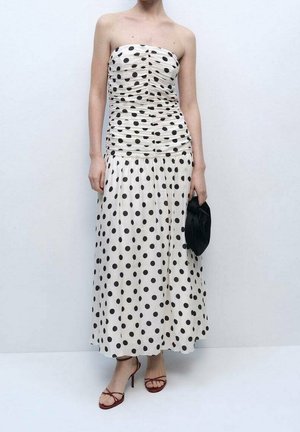 Femme portant une robe blanche bustier à pois noirs, tenant une petite pochette noire et portant des talons rouges à brides.