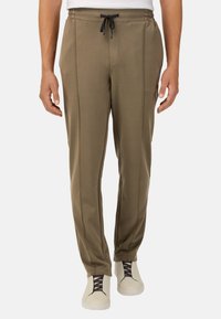 Alyvuogių žalios spalvos sportinės kelnės, pagamintos iš švelnaus audinio, turinčios elastingą juosmenį su raišteliu, šonines kišenes ir subtilius siūlus.