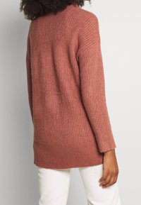 Rostfärgad stickad tröja med strukturerad yta, långa ärmar och avslappnad passform. I kombination med vita byxor, visad från baksidan.
