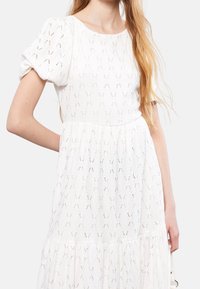 Kobieta w białej sukience z koronką eyelet, z rękawami bufiastymi i marszczonym pasem, z długimi blond włosami na jednolitym jasnym tle.