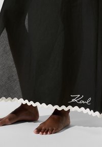 Sort gennemsigtigt stof med en flæset hvid kant, der har et diskret skriftmønster nederst. Blotte fødder står på en grå overflade.