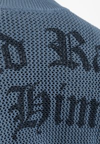 Világoskék kötött pulóver, textúrált hálós mintával, fekete betűzéssel, kontrasztos betűtípussal az anyagon.