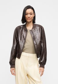 Brown leather bomber jacket with a shiny finish, ribbed cuffs, and collar. Paired with a beige top and light-colored trousers.