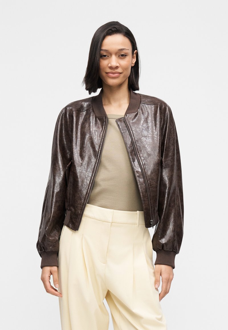 Brown leather bomber jacket with a shiny finish, ribbed cuffs, and collar. Paired with a beige top and light-colored trousers.
