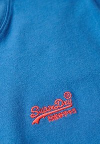 Blauwe katoen stof heeft een opvallend rood geborduurd logo "SuperDry" in de rechterbenedenhoek, met een zachte textuur en een gladde afwerking.