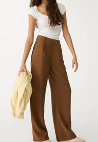 Brown wide-leg trousers with front pleats, paired with a white fitted top. Model holds a light yellow sweater in left hand. Smooth fabric texture.