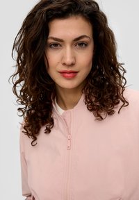 Veste bomber rose clair avec une fermeture éclair à l'avant, un col rond et une texture lisse. Des cheveux bouclés encadrent le visage, avec un maquillage naturel.