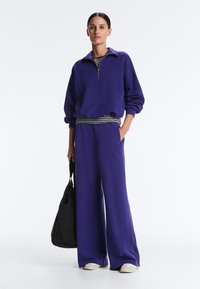 Kobieta stoi, ubrana w fioletową bluzę na suwak i szerokie spodnie, niesie czarną torbę, ma na nogach białe trampki i podkoszulek w paski.