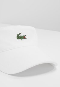 Hvit bomullshette med en buet skjerm som har en grønn krokodille-logo foran. Teksturen er glatt med et klassisk design.