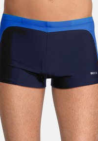 Shorts de bain navy avec une taille bleue et des accents latéraux. Matière lisse et extensible. Longueur de jambe courte, coupe ajustée, logo sur le côté.