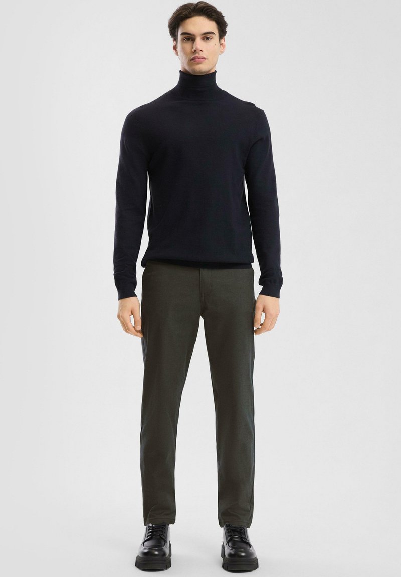 Maglione a collo alto blu navy, design aderente, abbinato a pantaloni verde oliva. Scarpe nere stringate con suola spessa completano il look.
