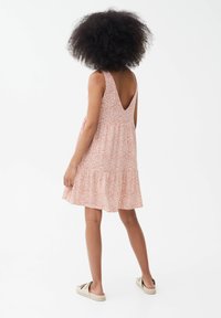 Robe sans manches à imprimé floral en rose et blanc, avec un dos en V profond et un ourlet superposé, fabriquée à partir d'un tissu léger. Des chaussures décontractées sont portées.