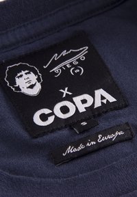 Zwarte stoffen label met witte tekst en een grafische illustratie. Bevat het woord "COPA," een handtekening en "Made in Europe."