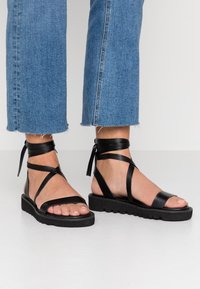 Svarta lädersandaler med ankelsnören och knyt detaljer. Tjock, texturerad sula med en lätt mönstring som kompletterar den minimalistiska designen.