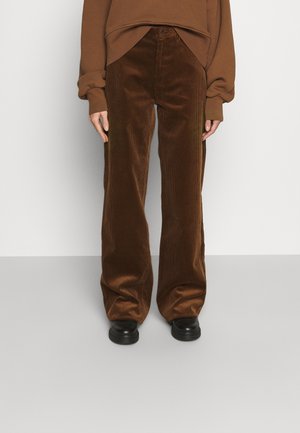 Person wearing loose brown corduroy pants, black shoes, and a matching brown long-sleeve top against a plain gray background.