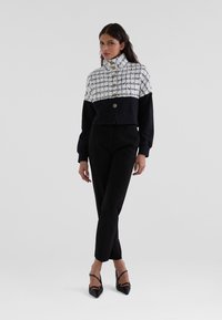 Cropped jacket in white and black pattern with high collar, featuring gold buttons. Paired with fitted black pants and black pointed shoes.