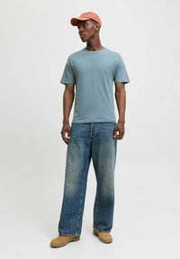 Light blue cotton t-shirt, relaxed fit, paired with faded wide-leg blue jeans, tan work boots, and a coral cap with embroidered detail.