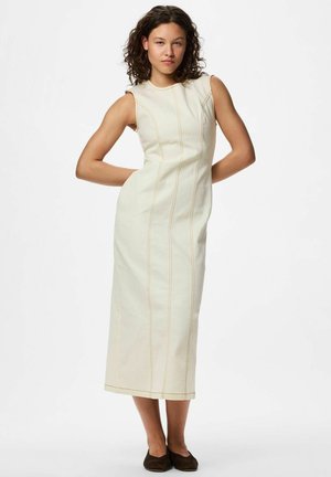 Mujer con cabello rizado que lleva un vestido sin mangas, color crema, hasta la pantorrilla con costuras verticales, de pie con las manos detrás de la espalda y zapatos planos negros.