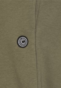 Olijfgroene t-shirt van een gestructureerde stof. Heeft een cirkelvormige zwart-wit patch met het label "Cars Jeans" aan de linkerkant.