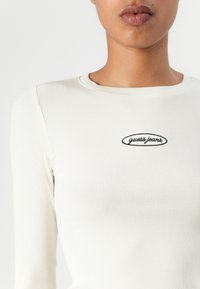 Dlouhý rukáv ribbed top v off-white barvě s kulatým výstřihem. Na přední straně se nachází malé vyšívané logo "guess jeans" v černé barvě.