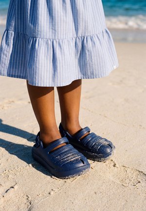 Kind draagt een blauw gestreepte jurk en donkerblauwe sandalen en staat op een zandstrand nabij het water.