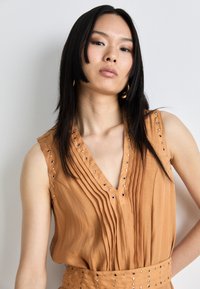 Haut sans manches en tissu beige chaud avec des plis verticaux et des œillets en métal décoratifs le long de l'encolure et des épaules.