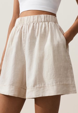 Nő bézs színű, magas derekú, rugalmas derékpánttal és oldalzsebekkel ellátott len rövidnadrágot visel, fehér crop toppal párosítva.