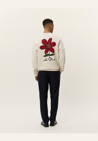 Sudadera color crema con un gran gráfico de flor roja en la espalda, con hojas verdes y contorno negro. El texto dice "Les Deux". Combinada con pantalones oscuros.