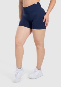 Marineblauwe hoge shorts, gemaakt van rekbaar materiaal, hebben een gladde textuur en een subtiele geribbelde tailleband. Gecombineerd met witte sportieve schoenen.