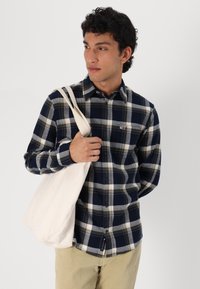 Navy and cream plaid shirt with button closure, long sleeves, and small logo detail. Model holds a beige canvas tote bag.