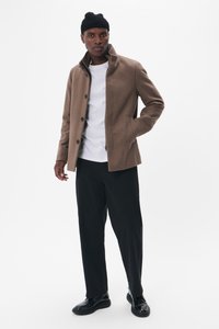 Chaqueta de mezcla de lana marrón con cuello alzado, cierre de botones y bolsillos laterales, combinada con una camiseta blanca y pantalones negros.