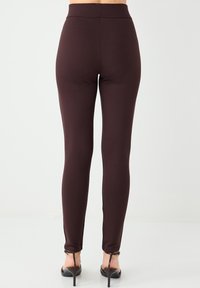 Brązowe legginsy z wysokim stanem, dopasowanym krojem i gładką fakturą. Noszone z czarnymi sandałami na wysokim obcasie. Widok z tyłu.