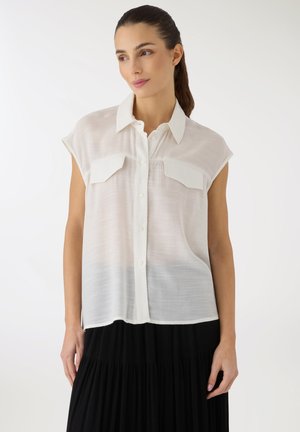 Donna con capelli scuri raccolti in una coda di cavallo, indossa una camicia bianca senza maniche abbottonata e una gonna nera plissettata su uno sfondo semplice.