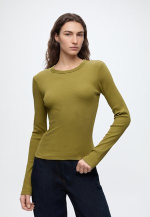 Femme aux longs cheveux ondulés portant un top ajusté à manches longues vert olive et un jean bleu foncé, debout devant un fond clair uni.