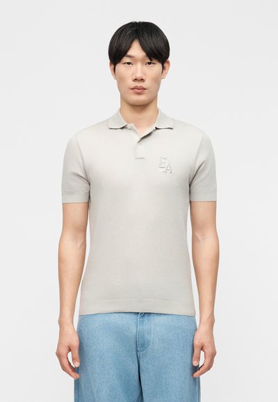 Jeune homme aux cheveux noirs portant un polo à manches courtes beige clair avec le logo "EA" et un jean en denim bleu clair, debout devant un fond blanc uni.