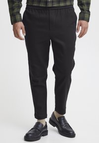 Svart bukser med elastisk midje og smal passform, laget av et glatt stoff. Brukt med svarte loafers og beige sokker, som viser et rent design.