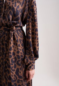 Abito con stampa leopardata marrone e nera, maniche lunghe, cintura legata in vita e bottoni davanti. Il tessuto appare liscio e lucido.