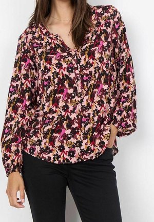 Mujer vistiendo una blusa de manga larga rosa con un patrón floral en rojo, negro y marrón, y pantalones negros, con la mano en el bolsillo.