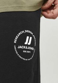Juodi medvilniniai laisvalaikio šortai su apvaliu baltu logotipu ir tekstu, esančiu šalia juosmens, po šviesiai žalia viršutine sluoksniu.