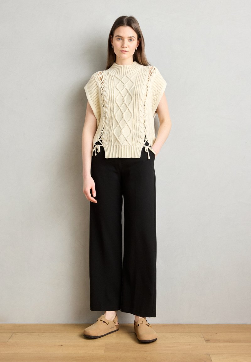 Maglione corto a maniche corte con lavorazione a treccia e lacci laterali. Abbinato a pantaloni neri a gamba larga e scarpe slip-on beige.